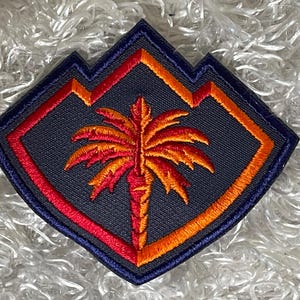 May include: Embroidered patch featuring a palm tree design in shades of orange and red, set against a dark gray background. The patch has a diamond shape with a blue border and orange accents, creating a vibrant and detailed emblem.