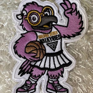 May include: Embroidered patch featuring a purple bird mascot wearing glasses, a basketball uniform, and holding a basketball. The patch has the words "GOLDEN STATE VALKYRIES" on the uniform. The patch is outlined in white.