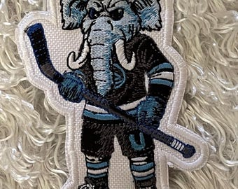 May include: Embroidered patch featuring an elephant mascot in a hockey uniform. The elephant is in black and blue, holding a hockey stick. The patch has a white border and is on a textured white background.