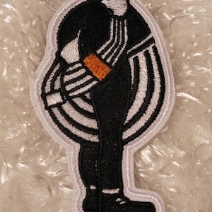 May include: Embroidered patch featuring a figure in a black and white hockey uniform, with a white outline. The figure is bent over, with the arms and back visible. The patch includes black skates and an orange accent.