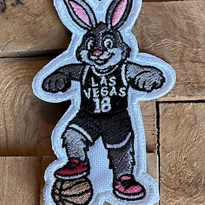 May include: Embroidered patch featuring a cartoon rabbit in a basketball uniform. The rabbit is wearing a black jersey with "LAS VEGAS 18" on it, black shorts, and red sneakers. The patch has a white border.