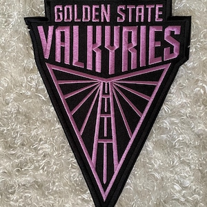 May include: A black and pink embroidered patch with the words "GOLDEN STATE VALKYRIES" in a stylized font. The patch has a triangular design with a geometric pattern and a black border.