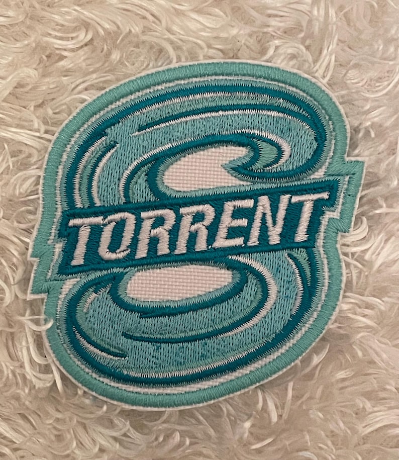 May include: Embroidered patch with the word "TORRENT" in white block letters. The patch features a stylized wave design in shades of teal and light blue, outlined in white and teal. The patch is on a white, fluffy surface.