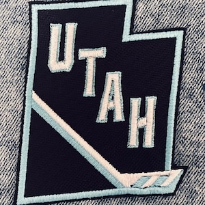 May include: Embroidered patch in the shape of Utah, featuring the word "UTAH" in white lettering. The patch has a navy blue background with light blue and white accents, including a hockey stick design.