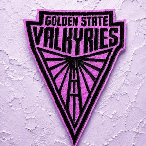 May include: A triangular embroidered patch featuring a vibrant pink background with bold black stitching. The text GOLDEN STATE VALKYRIES is prominently displayed at the top, while the lower section contains a black geometric sunburst design.