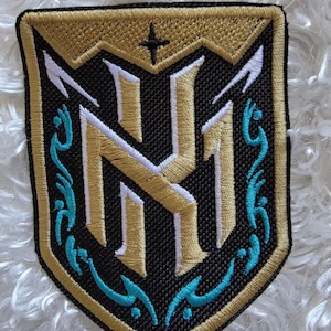 May include: A gold and white embroidered patch with a shield shape and a stylized "NH" logo in the center. The shield is surrounded by a turquoise blue border with a decorative design.