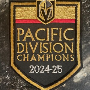 May include: A black and gold embroidered patch with the text "Pacific Division Champions 2024-25". A silver knight's helmet with a "V" is above the text.