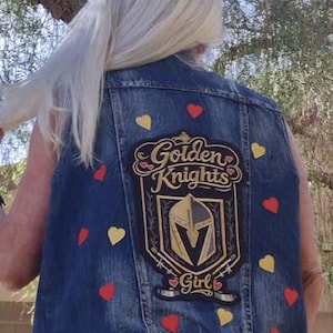 May include: A blue denim vest featuring a large rectangular patch on the back with the text Golden Knights Girl, a central helmet emblem, and crossed hockey sticks. The vest is decorated with scattered small red and yellow heart-shaped appliques.