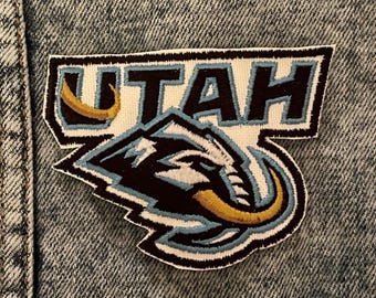 May include: Embroidered patch featuring the word "UTAH" in navy blue block letters above a stylized emblem. The emblem depicts a mammoth with light blue and gold accents, outlined in black and white. The patch is sewn onto a denim jacket.