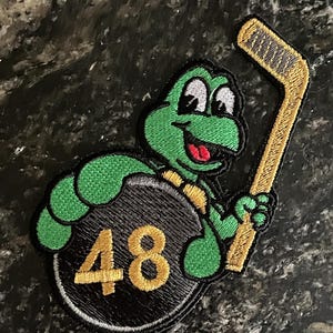 May include: Green turtle cartoon character holding a gold hockey stick and a black puck with the number 48 on it.