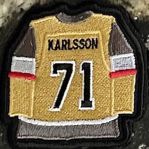 May include: A gold hockey jersey patch with black and gray accents. The jersey has the name "KARLSSON" and the number "71" on the back.