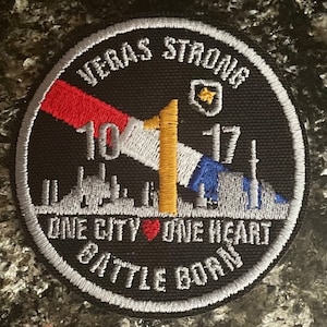 May include: A black and white embroidered patch with the text "Vegas Strong 10.1.17 One City One Heart Battle Born". The patch features a red, white, and blue American flag with a yellow star in the upper right corner.