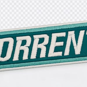 May include: Embroidered patch with the word "TORRENT" in white, block letters. The text is set against a teal background, framed by a teal border and a white outline. The patch has a rectangular shape with angled edges.