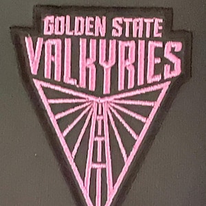 May include: A pink and black patch featuring the text "GOLDEN STATE VALKYRIES" above a pink triangle design. The triangle contains a road design. The patch is likely for apparel or accessories.