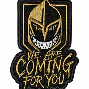 May include: An embroidered patch featuring a black and gold knight helmet with a wide, toothy grin. Below the helmet, the text WE ARE COMING FOR YOU is stitched in gold thread against a black background, with a shield-shaped border surrounding it.