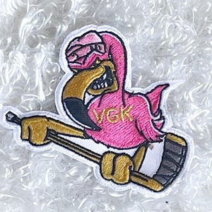 May include: A pink flamingo wearing a white hat and holding a hockey stick. The flamingo has a black beak and eyes, and the words "VGK" are written in gold on its chest.