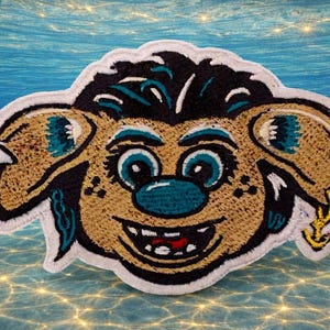 May include: Embroidered patch featuring a cartoonish face with a wide grin, large eyes, and a teal nose. The patch has a tan and black color scheme with teal and yellow accents, and a white border. The background is an underwater scene.