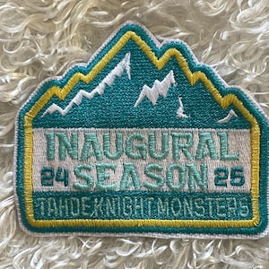 May include: Embroidered patch with a teal mountain range design, outlined in yellow and teal. The patch reads "INAUGURAL 24 SEASON 25 TAHOE KNIGHT MONSTERS" in white and teal lettering. The patch is rectangular with a mountain-shaped top.