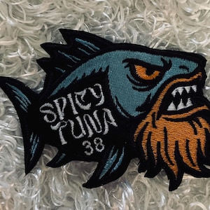 May include: Embroidered patch featuring a stylized fish with a fierce expression. The fish has a teal body, orange beard, and white teeth. The text "SPICY TUNA 38" is embroidered in white on a black background. The patch is likely for clothing or accessories.
