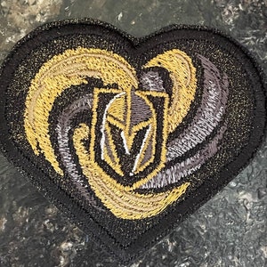 May include: A black felt heart-shaped patch with an embroidered gold and silver design. The design features a knight's helmet with a plume and a swirling pattern.
