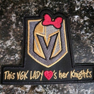 May include: Black embroidered patch with a gold and gray Vegas Golden Knights logo. A red bow is on top of the logo. The text "This VGK Lady's her Knights" is on a black banner below the logo.
