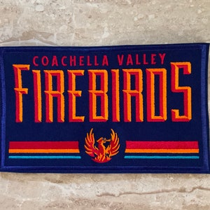 May include: A navy blue embroidered patch featuring the words "COACHELLA VALLEY FIREBIRDS" in orange and red lettering. Below the text is a phoenix emblem and horizontal stripes in red, orange, and blue.