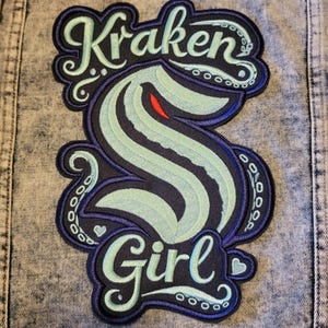 May include: Close-up of a denim jacket back with a large embroidered patch. The patch features the words "Kraken Girl" in a stylized font, with a teal and navy blue kraken design. The jacket has a light blue wash.
