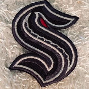 May include: Embroidered patch in the shape of a stylized letter "S". The patch features a navy blue background with white and gray stripes, and a red accent. The edges are outlined with white stitching.
