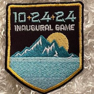 May include: A black patch with a gold border featuring the text "10+24+24 INAUGURAL GAME". The patch has a teal and white mountain range over a teal body of water, with a yellow sun in the background.