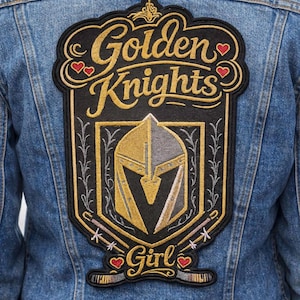 May include: A denim jacket with a large, embroidered patch on the back. The patch features the words "Golden Knights" in gold lettering, a knight's helmet, and the word "Girl" in a decorative font. The patch is outlined in black.