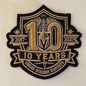 May include: A black and gold embroidered patch featuring the number 10 with a hockey trophy and a knight helmet. The text reads 2017, 2027, 10 YEARS, and VEGAS GOLDEN KNIGHTS. The design is shaped like a shield with decorative banners and star accents.