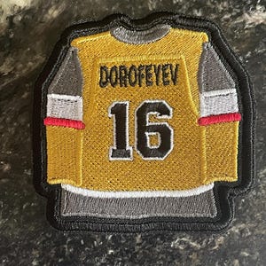 May include: A yellow and gray embroidered patch of a hockey jersey with the name "DOROFEEYEV" and the number "16" on the back.