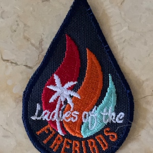 May include: A blue, teardrop-shaped patch with an orange and red flame design. A white palm tree is in the center of the flame. The text "Ladies of the Firebirds" is embroidered in white on the patch.
