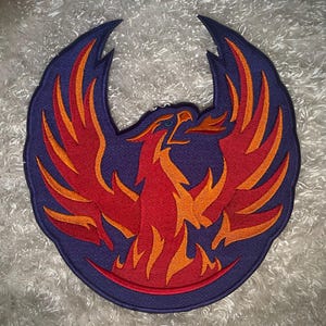 May include: A circular patch featuring a phoenix design. The phoenix is depicted in shades of red and orange, with a blue outline and background. The patch has a detailed embroidered design.