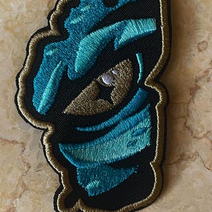 May include: A teal blue and gold embroidered patch with a stylized design of a face wearing a mask.
