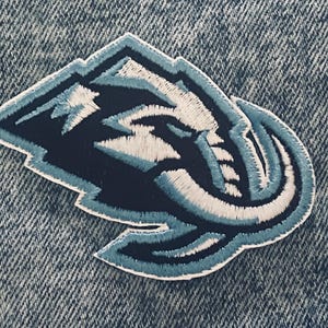 May include: Embroidered patch featuring a stylized elephant head within a mountain-shaped outline. The patch uses shades of blue, white, and navy, with detailed stitching. The patch is likely designed for apparel or accessories.