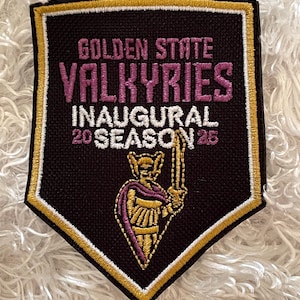 May include: A black and gold patch with the text "GOLDEN STATE VALKYRIES INAUGURAL 20 SEASON 25". The patch features a gold border and a purple and gold illustration of a warrior holding a sword.