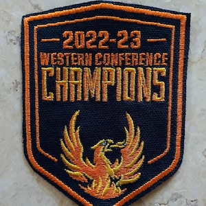 May include: Embroidered patch with a shield shape, featuring the text "2022-23 Western Conference Champions" in orange. A stylized orange phoenix is at the bottom. The patch has a black background with orange trim.