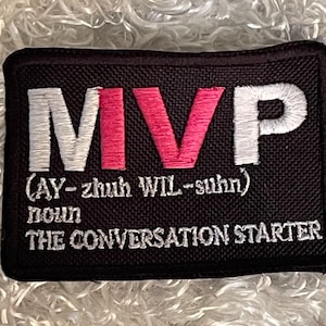 May include: Black rectangular patch with white and pink embroidered text. The text reads "MVP" with the phonetic pronunciation below. Below the pronunciation, the text reads "noun THE CONVERSATION STARTER".