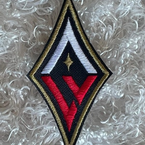 May include: A diamond-shaped patch with a black background, featuring white and red geometric designs, and a gold border. A small gold star is centered above the red design. The patch is likely for clothing or accessories.