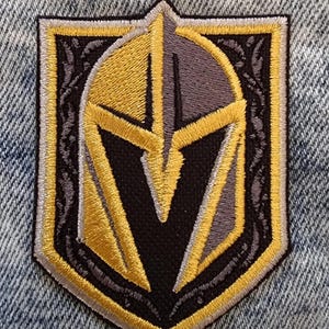 May include: Embroidered patch featuring the Vegas Golden Knights team logo. The patch has a black, gold, and gray color scheme, with a shield shape and intricate stitching details. The patch is set against a denim background.