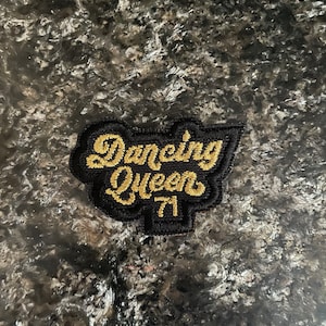 May include: A black and gold embroidered patch with the text "Dancing Queen 71" in a cursive font.