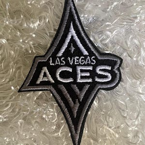 May include: Embroidered patch in a diamond shape with the words "LAS VEGAS ACES" in white lettering. The patch has a black background with silver and white accents. The patch is likely for a sports team or fan apparel.