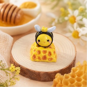 May include: A small, handcrafted artisan keycap featuring a yellow bee with a tiny crown, black stripes, and light blue wings, resting atop a yellow base with a textured, honeycomb-inspired pattern in shades of orange and yellow.