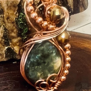 May include: Handmade copper wire pendant with a large, oval, green gemstone at the center. The pendant features intricate wire wrapping, small copper beads, and a gold-colored bead. The pendant is displayed on a wooden surface.