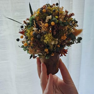 Peut inclure: Un petit bouquet rond de fleurs et de feuillages séchés dans un pot miniature marron. L'arrangement présente un mélange de baies jaunes, vertes, blanches et bleu foncé, ainsi que diverses tiges séchées, herbes et petites fleurs aux tons terre.