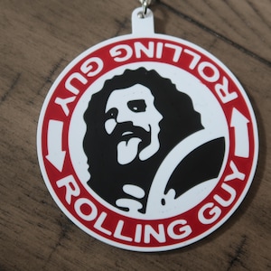 May include: A white and red circular keychain with the words "Rolling Guy" and an illustration of a bearded man with his tongue out. The keychain is hanging on a wooden surface.
