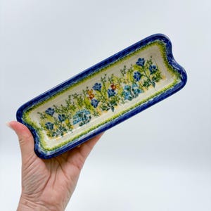 May include: A rectangular ceramic tray with a deep blue border and a hand-painted interior featuring green foliage, small blue flowers, and yellow bees. The tray has a slightly curved, indented rim design and a smooth, glossy finish throughout.