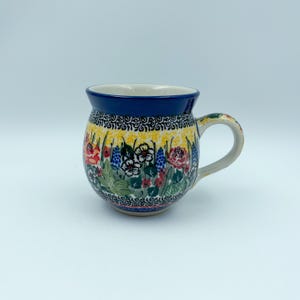 May include: A round ceramic mug featuring a dark blue rim and a handle. The body is decorated with a vibrant floral pattern including red, yellow, and blue flowers, green leaves, and black scrollwork details against a light yellow background.