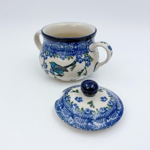 May include: A ceramic sugar bowl with a lid, featuring a blue bird and floral design. The bowl has two handles and a dark blue rim. The lid is decorated with blue flowers and a dark blue knob.